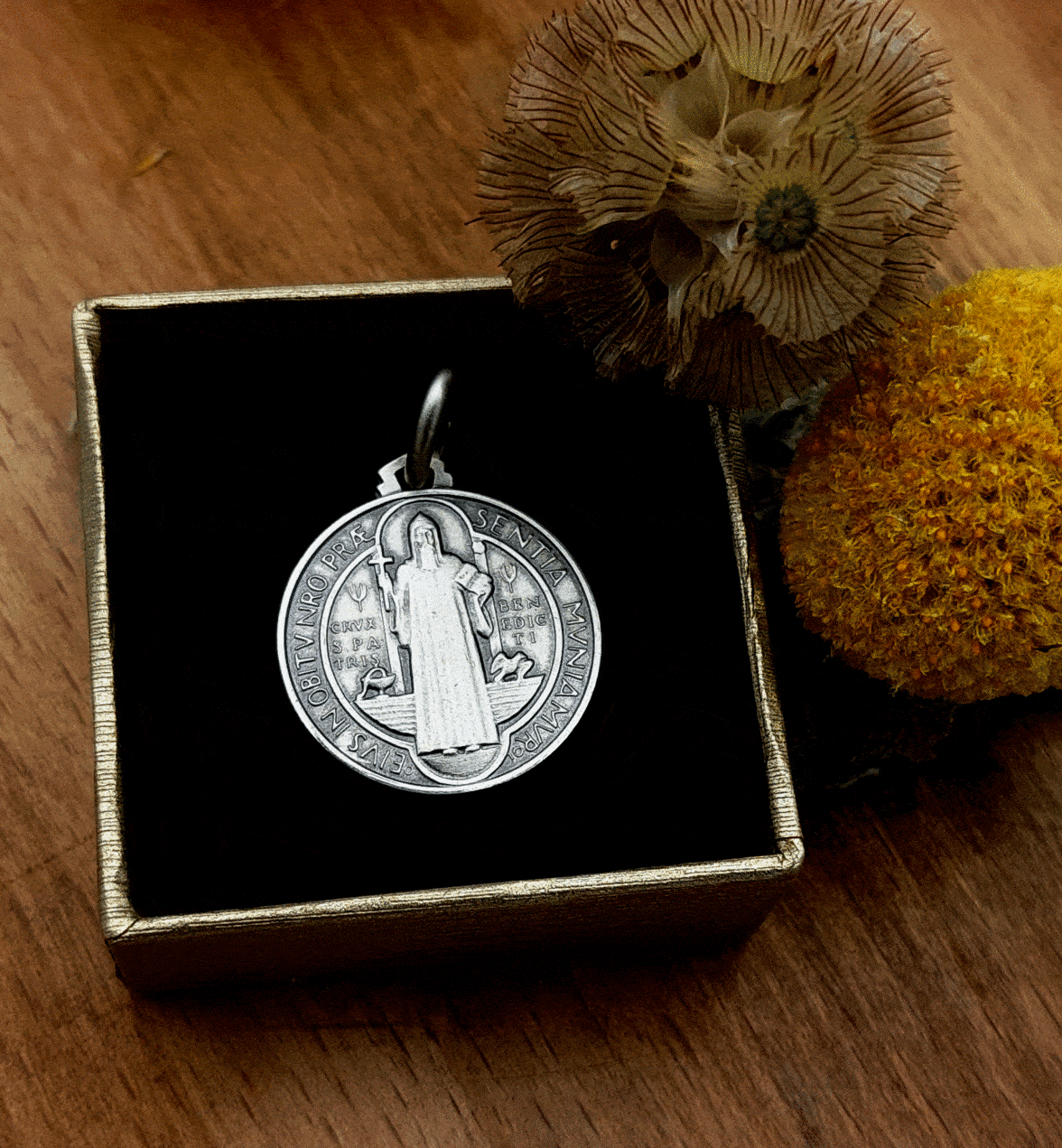 medalla con representación de san benito vestido con habito y sosteniendo una cruz y un libro y guardada en una caja y la caja rodeada de flores